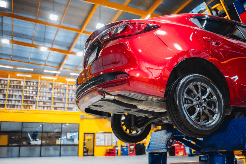 Extended Warranty Auto Repairs, auto repair in Seekonk, MA at Trustworthy Auto. Image of a red car lifted on a hydraulic automotive lift inside a brightly lit repair shop, showing the vehicle’s undercarriage and wheels as technicians prepare for maintenance or inspection.