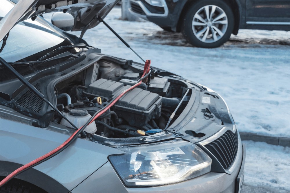 Battery, Starter, or Alternator, auto repair in Seekonk, MA at Trustworthy Auto. A car in a snowy parking lot with jumper cables attached to the battery, highlighting cold weather challenges and the need for vehicle preparedness.