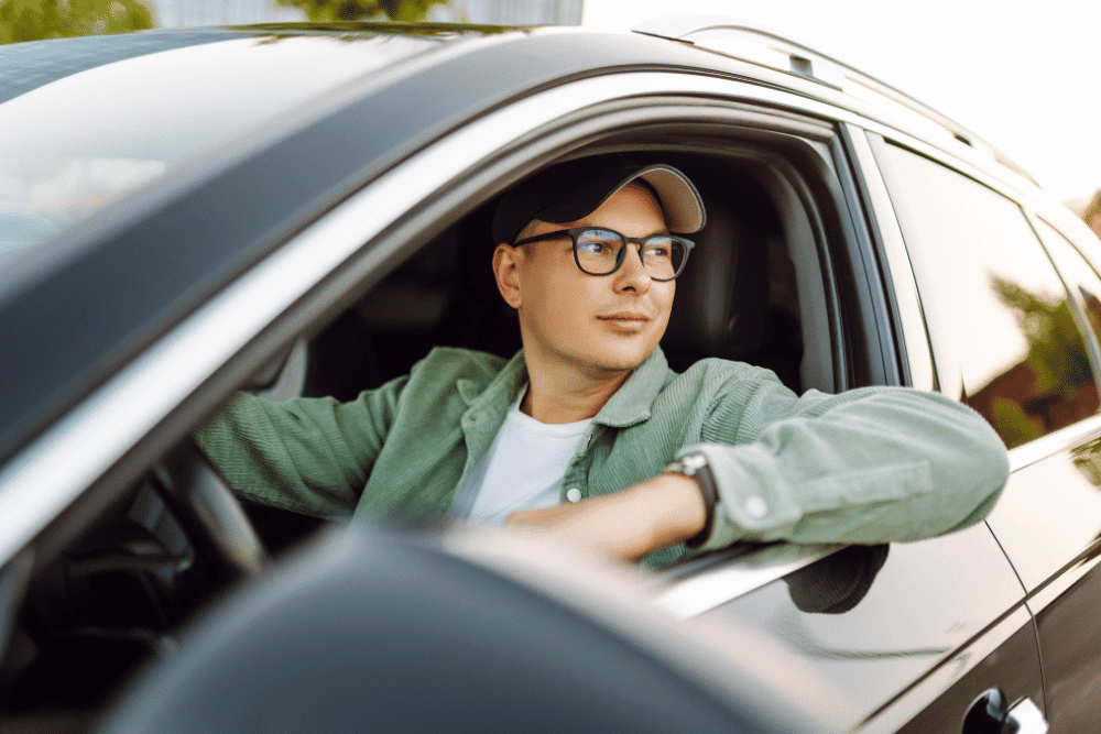 Summer Car Care Checklist for Massachusetts Drivers by Trustworthy Auto -- A man wearing glasses and a green jacket sits in a car, looking out the window