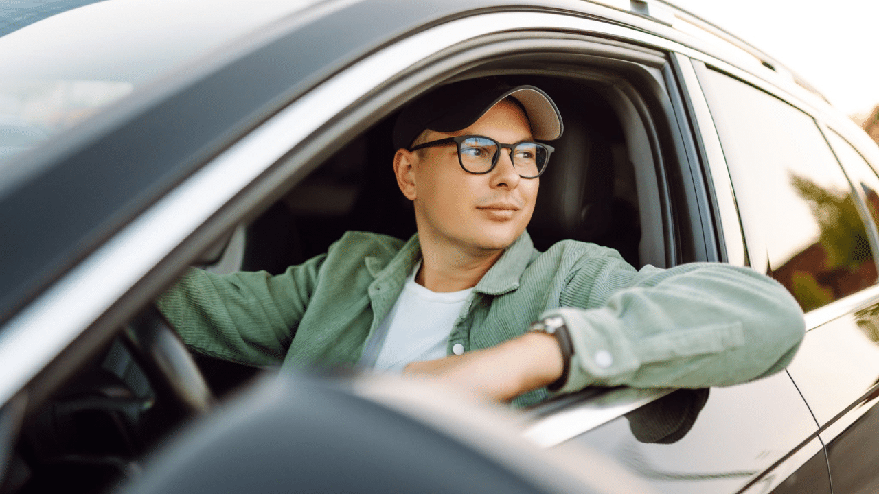 Summer Car Care Checklist for Massachusetts Drivers -- A man wearing glasses and a green jacket sits in a car, looking out the window with a relaxed and thoughtful expression. The sun sets in the background.
